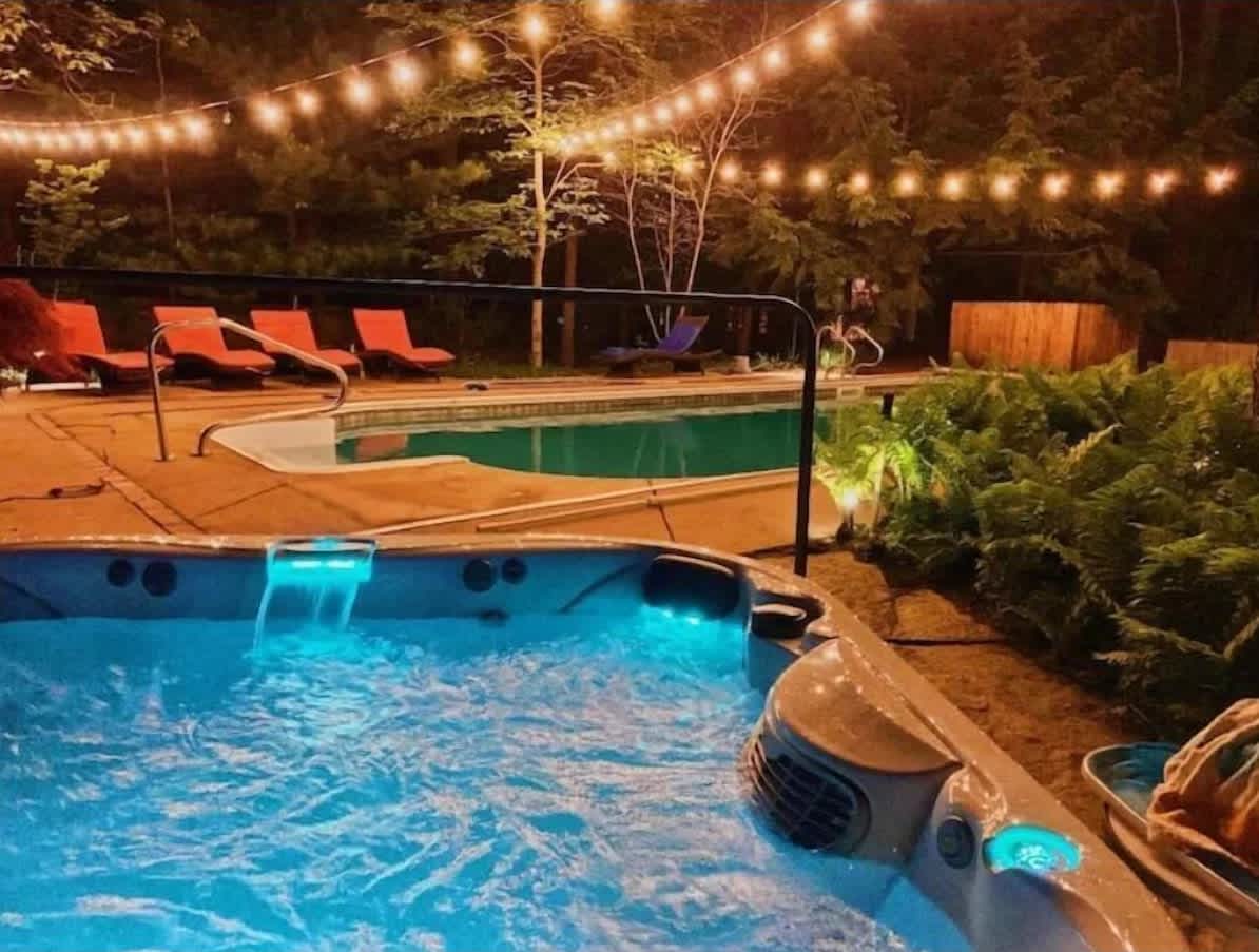 Nighttime view of a bubbling hot tub with blue LED lighting and waterfalls, next to a swimming pool illuminated by string lights overhead. Orange lounge chairs are visible by the pool, creating a cozy evening ambiance.