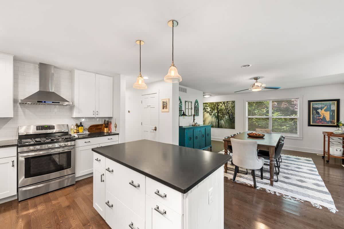 Well-appointed kitchen with a central island, white cabinetry, and stainless steel appliances, including a gas range and range hood. The space opens directly to the dining area with a wooden table and a teal buffet cabinet, and an adjacent entryway.