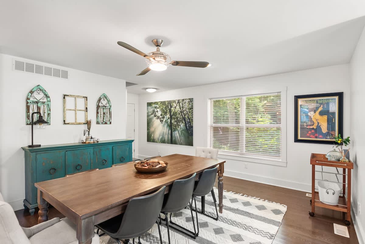Bright dining room showcasing a rustic wooden table and chairs on a geometric rug, with a teal buffet cabinet and decorative window frames on the wall. A large nature print and a vintage-style bar cart add character to the space.