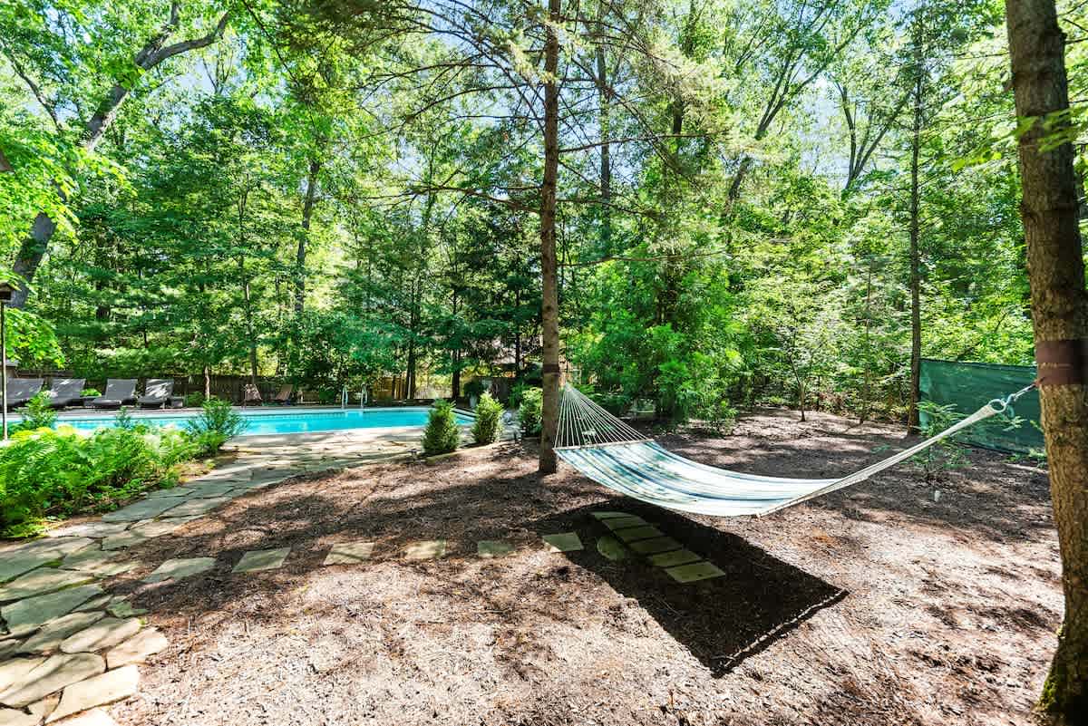 A striped hammock hangs between two tall trees in a shaded, wooded area with a dirt and mulch ground cover. A flagstone path leads towards a swimming pool and lounge chairs visible in the background, surrounded by dense foliage.