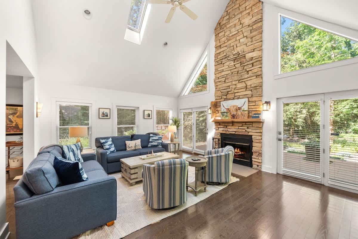 Spacious living room with a towering stone fireplace extending to the vaulted ceiling, flanked by large windows offering views of lush greenery. Blue sofas and striped armchairs are arranged around a rustic coffee table on a light area rug.