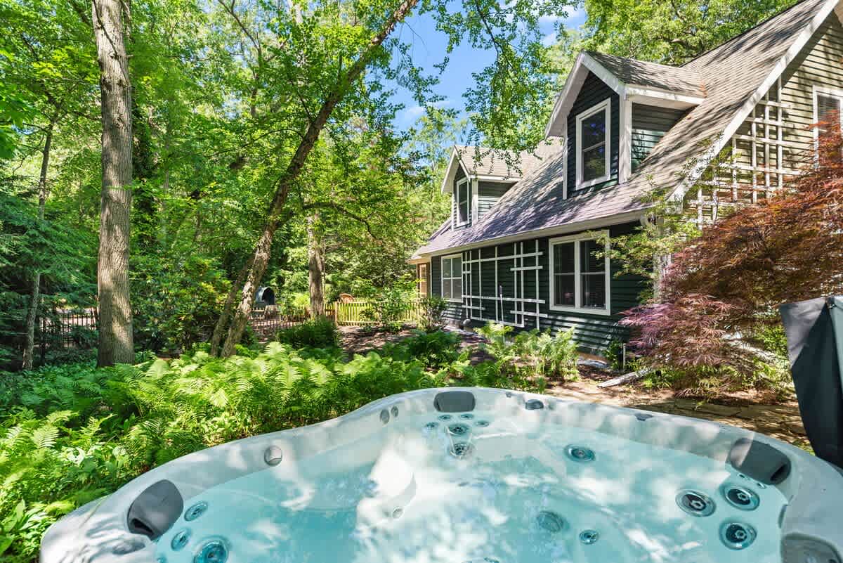 Close-up view of a bubbling outdoor hot tub with its cover partially open, set on a stone patio surrounded by lush ferns and greenery, with the dark gray house exterior visible in the background.