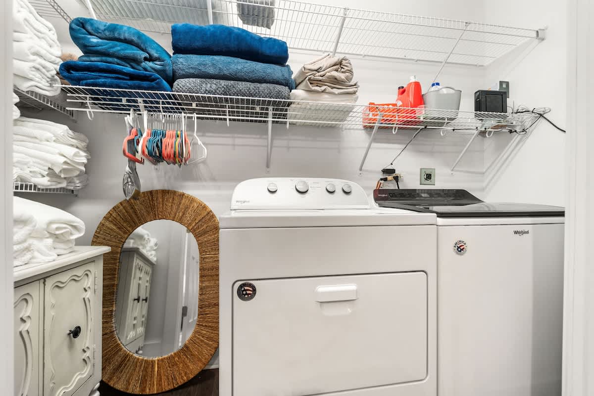 Functional laundry room equipped with a white washer and dryer, wire shelving for storage, and a large round mirror. Various towels and laundry supplies are neatly organized.