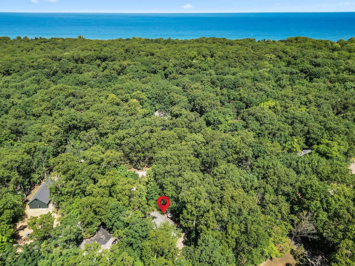 High aerial view showcasing the vacation rental property nestled within a vast, dense green forest, with the blue expanse of a large body of water (likely Lake Michigan) visible in the distance. A red pin marks the property's location.