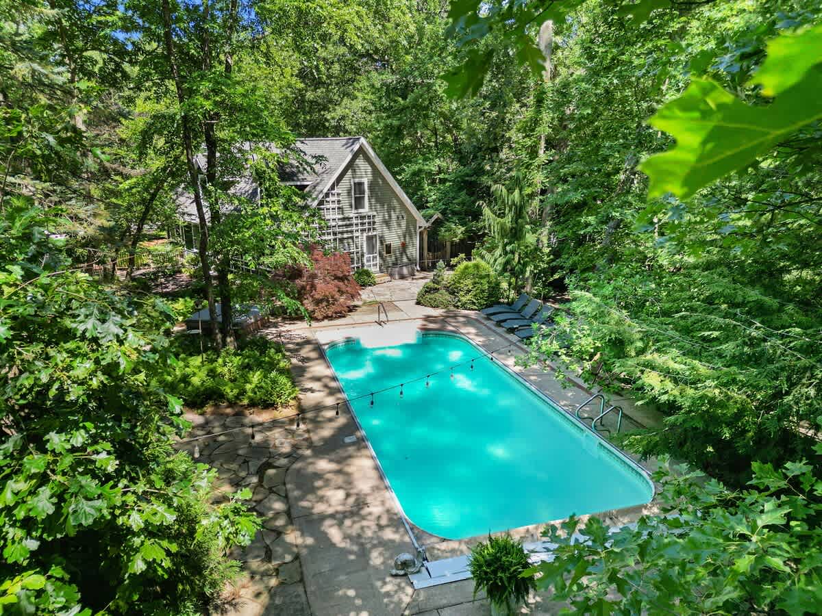 Aerial view of a gray-sided house with dormer windows, a large rectangular swimming pool on a stone patio, and dense green trees surrounding the entire property under a bright sky.