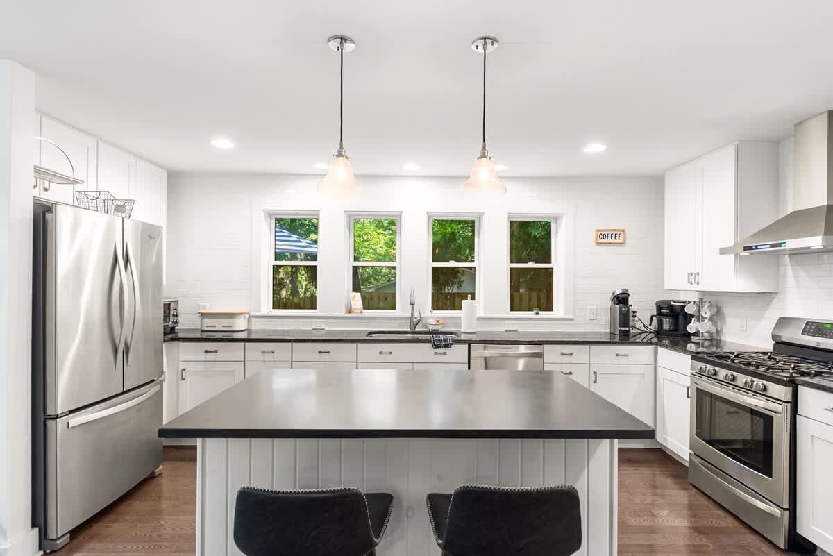 Modern kitchen with white shaker cabinets, dark countertops, stainless steel appliances, and a large island with two bar stools. Three windows above the sink provide natural light.