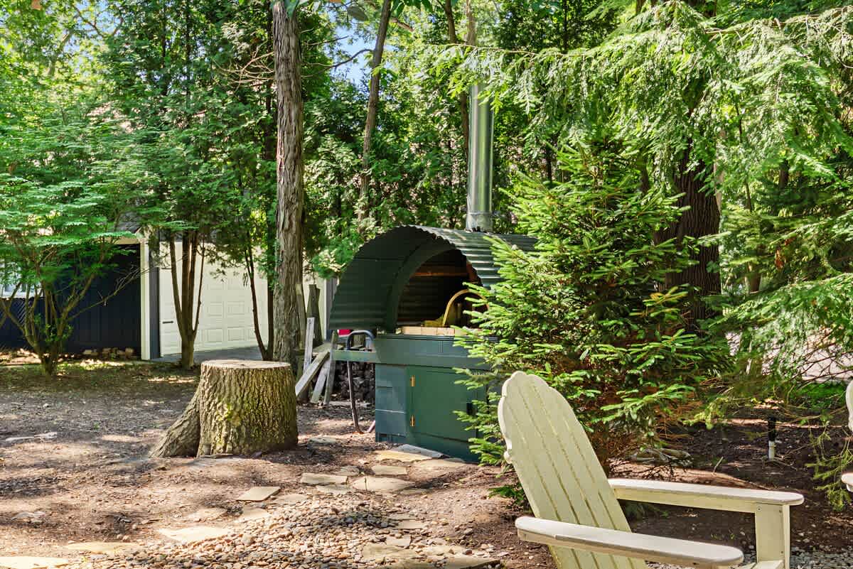 Outdoor pizza oven with a green base and a corrugated metal dome, situated on a gravel patch next to a large tree stump and surrounded by dense green foliage, with a white Adirondack chair in the foreground.