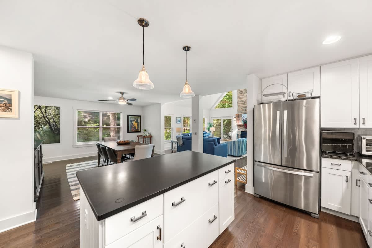 Open-concept kitchen with a large island and stainless steel French door refrigerator, offering a clear view into the dining area and beyond to the living room. White cabinets and black countertops maintain a sleek, modern aesthetic.