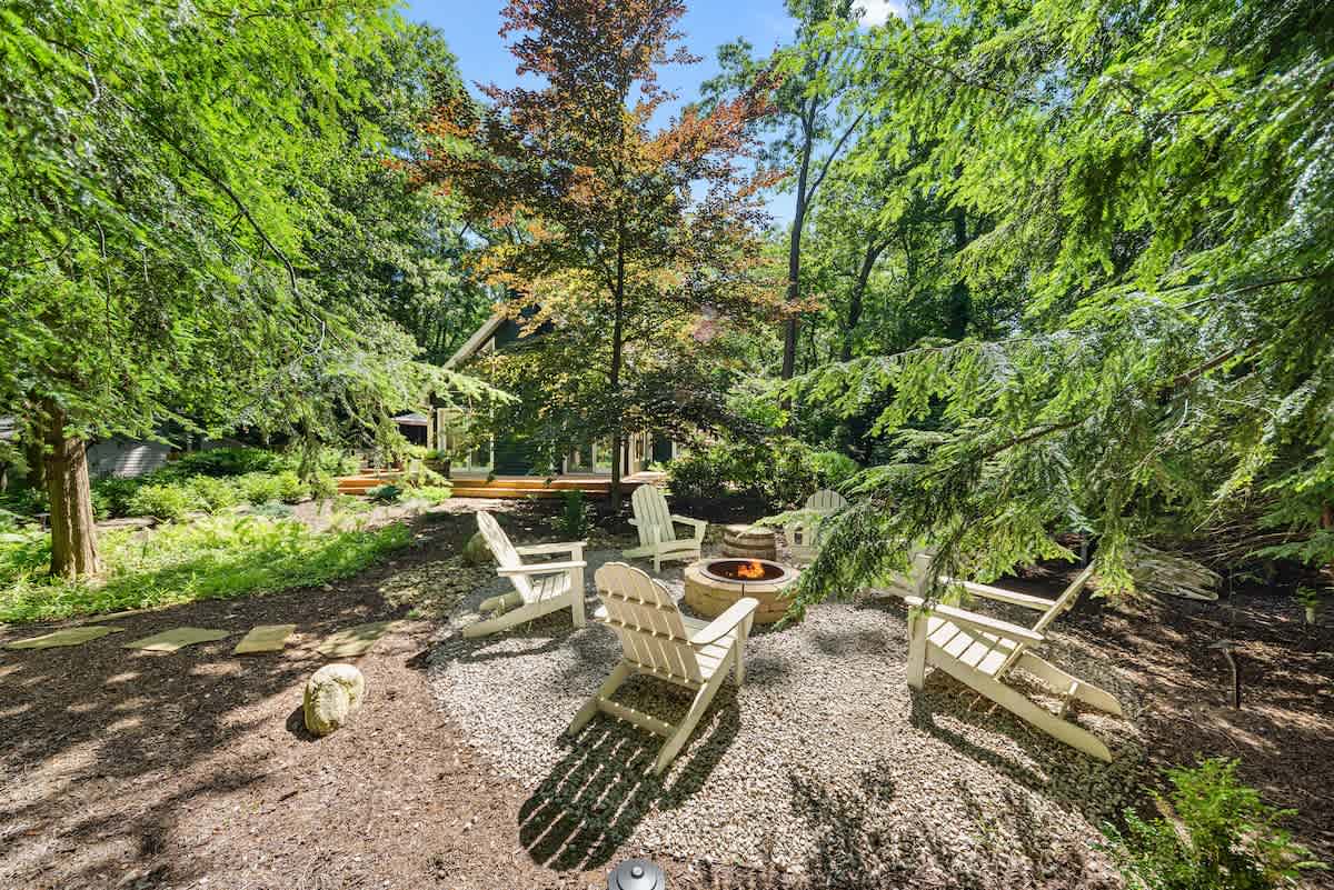 Outdoor fire pit area with a circular gravel base, surrounded by four white Adirondack chairs, nestled among lush green trees and foliage, creating a secluded and natural setting.