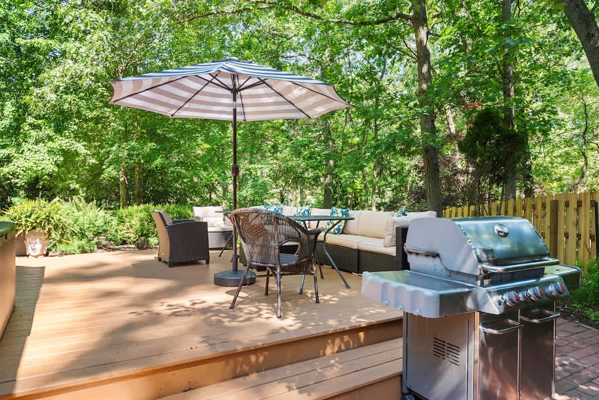 Wooden deck with a stainless steel BBQ grill in the foreground, an outdoor dining set with an umbrella, and a large sectional sofa in the background, all set amidst a backdrop of dense green trees.