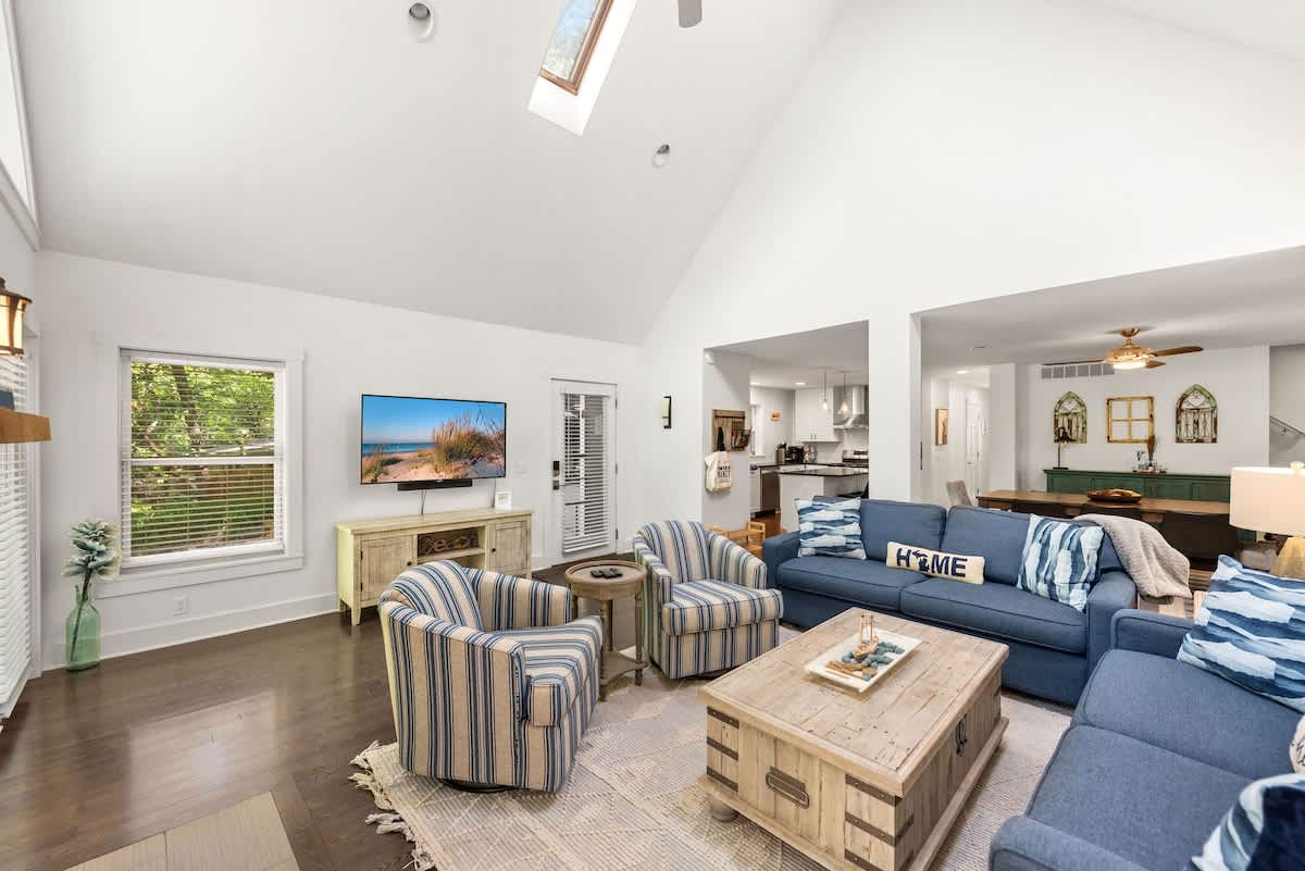 Cozy living room featuring a wall-mounted TV above a light wood console, with two blue sofas and two striped armchairs centered around a wooden coffee table. Dark hardwood floors and white walls create a clean, inviting space.