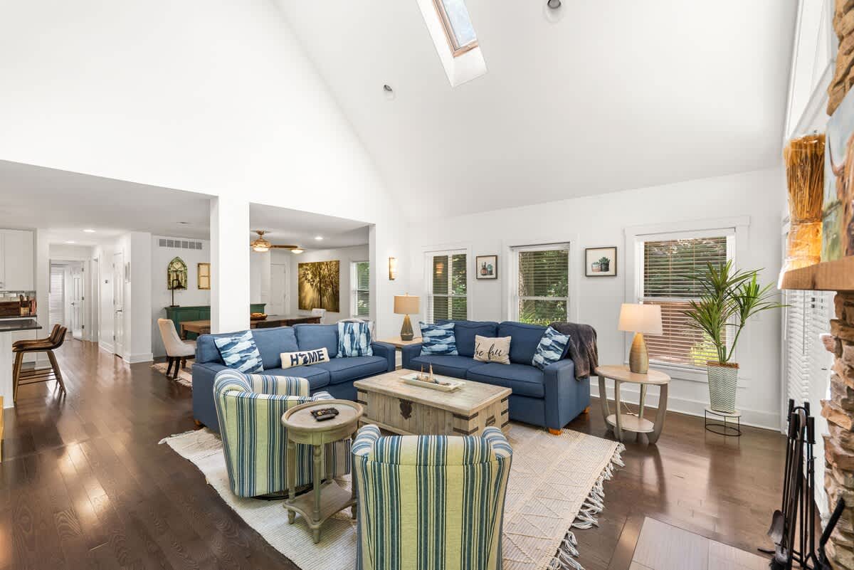 Bright and airy living room with vaulted ceilings and skylights, featuring two blue sofas, two striped armchairs, a rustic wooden coffee table, and dark hardwood floors. An open layout connects to the dining area and kitchen beyond.