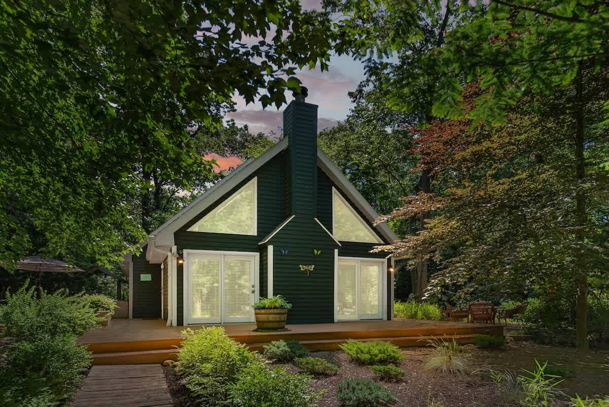 Evening view of the dark green A-frame house with a tall chimney and large windows, framed by lush trees and foliage under a twilight sky. The wooden deck in front features outdoor seating, illuminated by soft lighting.