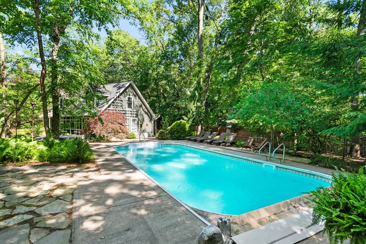 Ground-level view of a rectangular swimming pool with clear blue water, surrounded by a flagstone patio and lush green ferns. A dark green house with white trim is visible in the background, framed by tall trees.