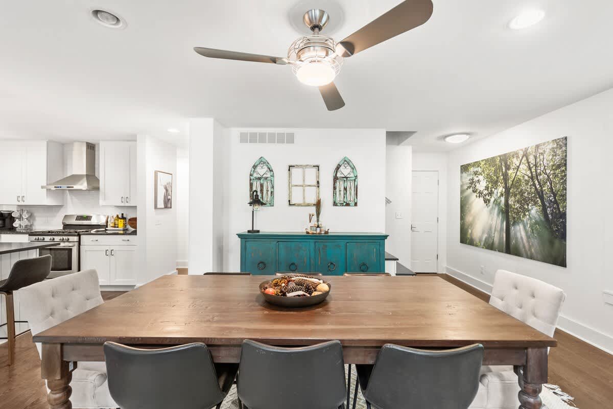 Elegant dining room with a large wooden table surrounded by a mix of upholstered and modern chairs, illuminated by a ceiling fan. A teal buffet cabinet with decorative windows and a large nature-themed artwork adorn the wall, with the kitchen visible in the background.