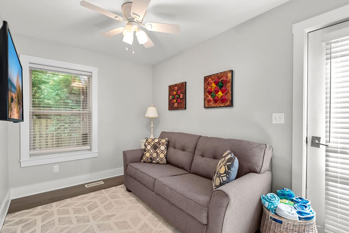 Bright living space with light gray walls, a gray sofa, and a wall-mounted TV. A window provides natural light, and a ceiling fan is installed overhead.