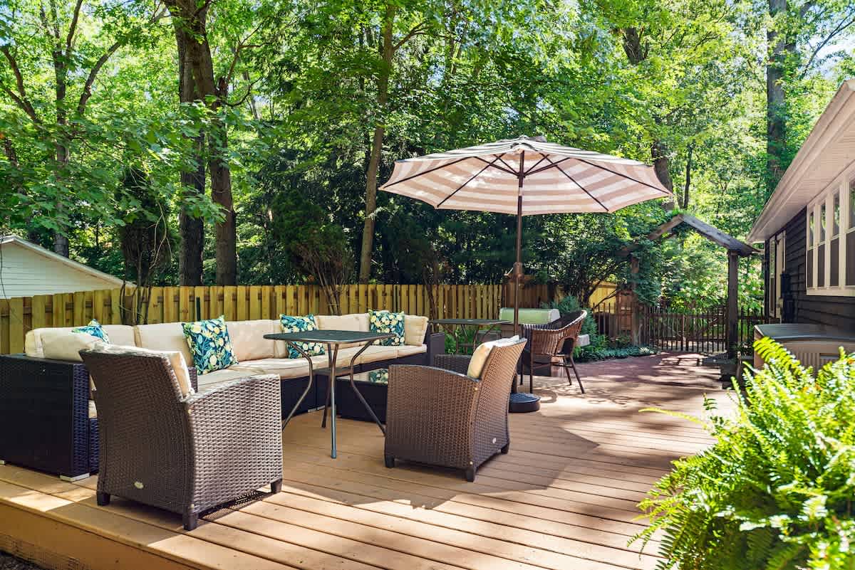 Spacious wooden deck featuring a large outdoor sectional sofa with light cushions, a dining table with chairs, and a striped umbrella, all surrounded by a wooden fence and lush green trees.