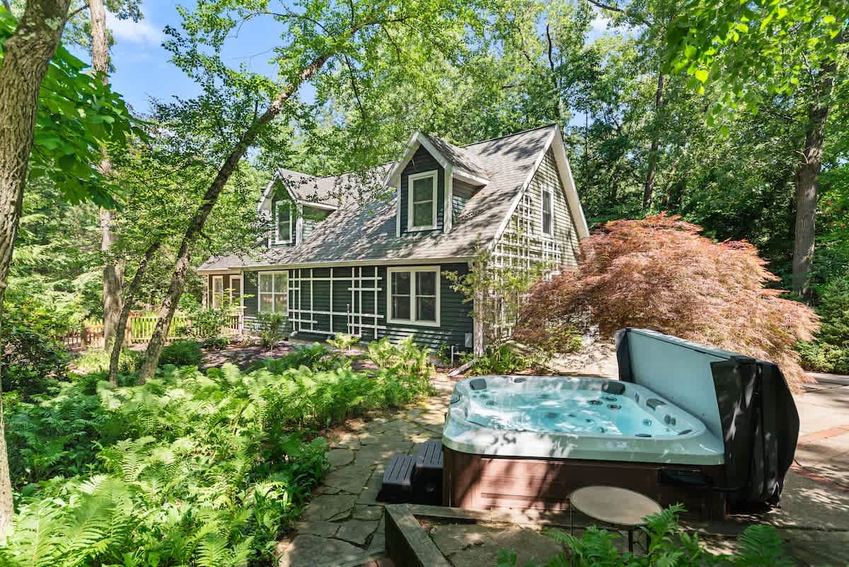 Exterior view of a dark gray house with white trim and dormer windows, surrounded by dense green trees and ferns, with an outdoor hot tub on a stone patio in the foreground.