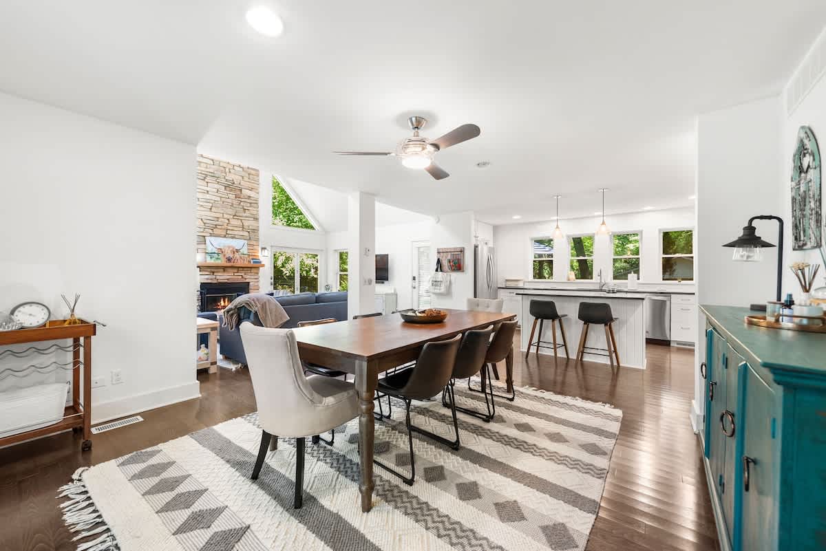 Open-concept dining area featuring a long wooden table with mixed seating on a patterned rug, under a ceiling fan. The space flows into the living room with a stone fireplace and the modern kitchen with an island and bar stools.