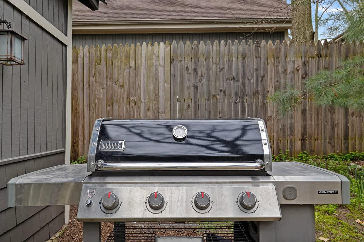 A stainless steel Weber Genesis II gas grill with a black lid sits on a paved surface next to the dark gray house, with a wooden fence in the background.