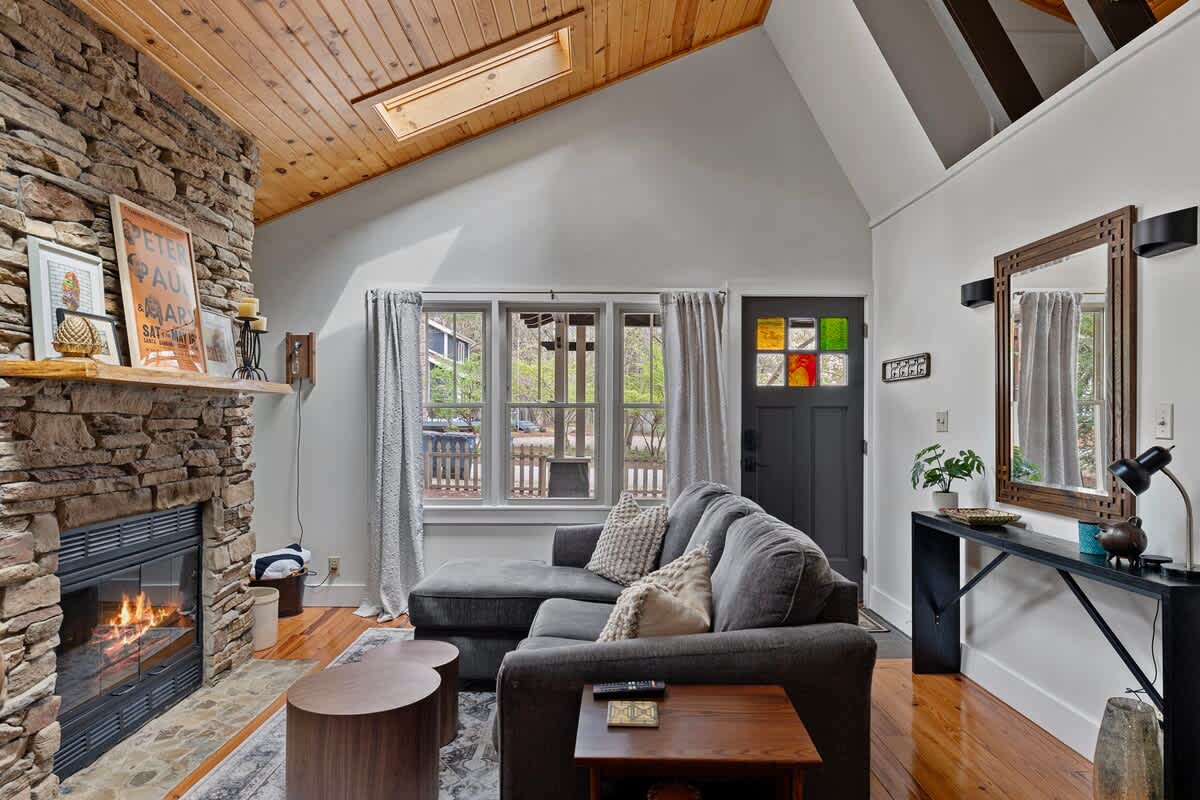 A bright living room showcases a gray sectional sofa, a large stone fireplace, and a dark front door with colorful stained glass accents, all under a high vaulted wooden ceiling.