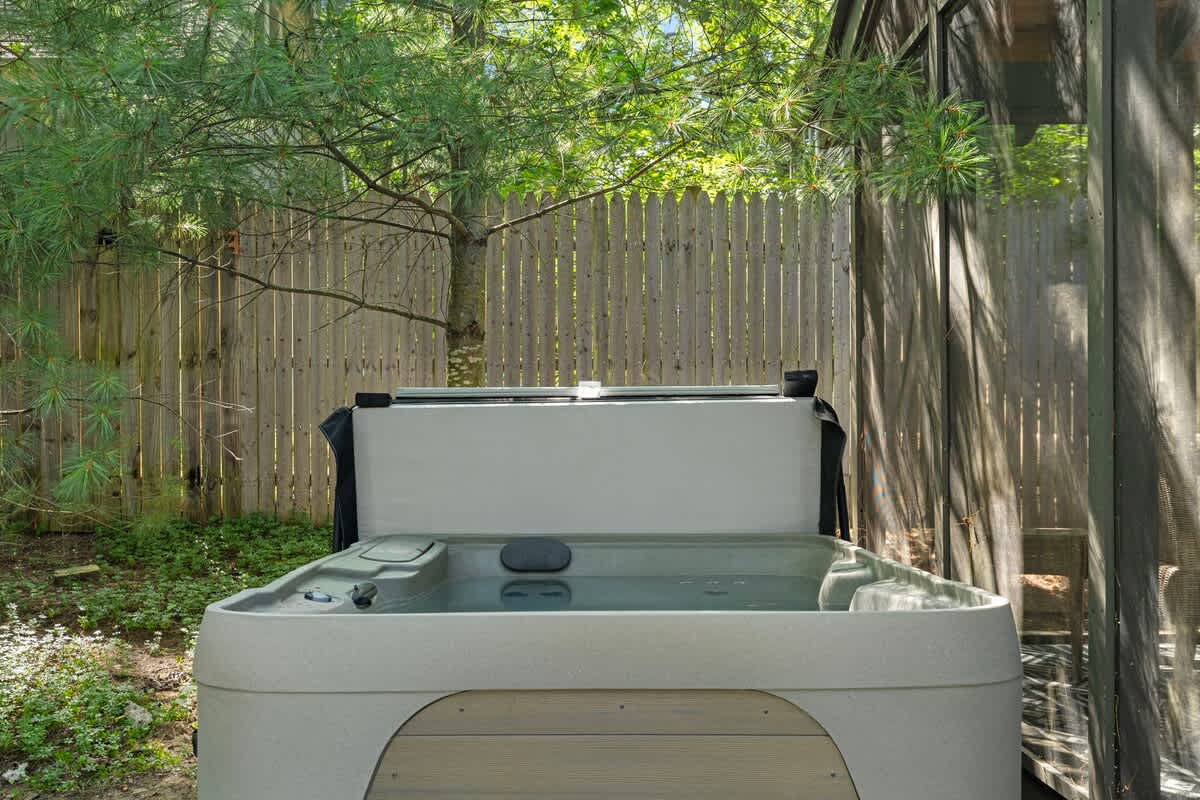 Outdoor hot tub with its cover partially open, nestled in a private area surrounded by a wooden fence and lush greenery. A screened structure is visible to the right.