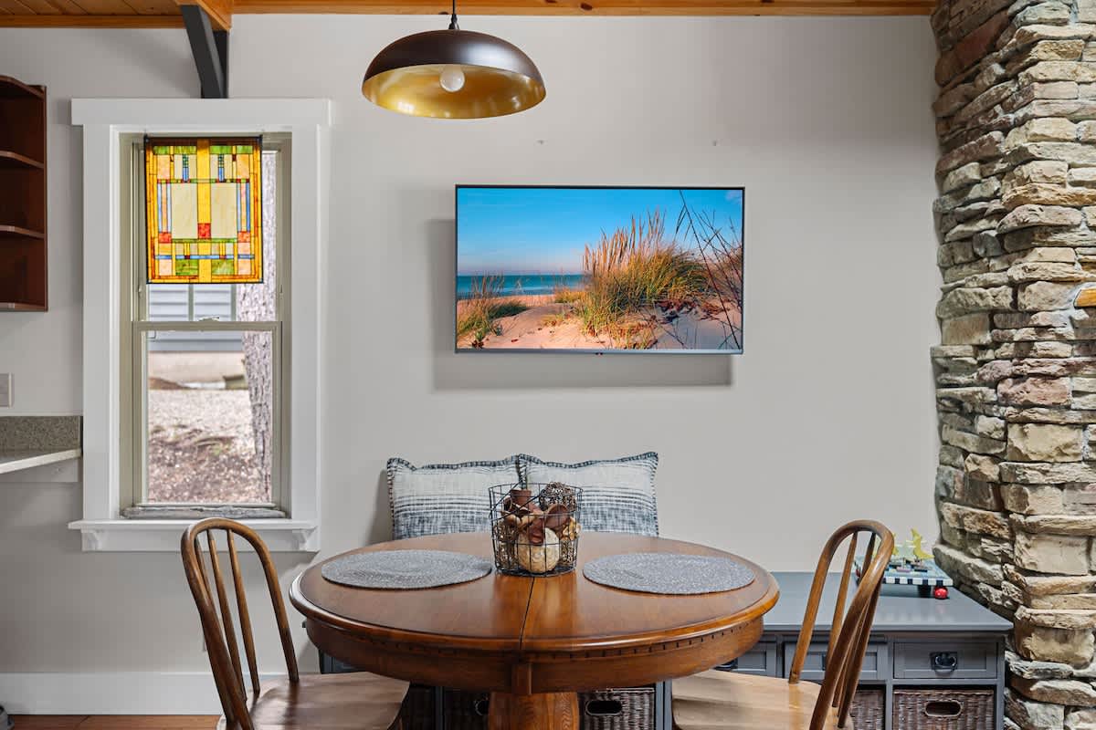 Cozy dining area featuring a round wooden table with two chairs, a wall-mounted TV displaying a beach scene, and a decorative stained glass window, all under a modern pendant light.