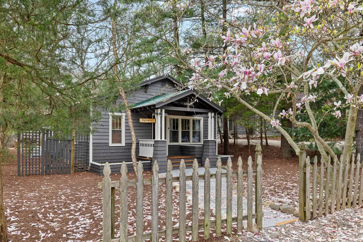 The charming front exterior of the dark gray house, framed by a blooming magnolia tree and a rustic picket fence. Pink petals are scattered on the mulch-covered ground.