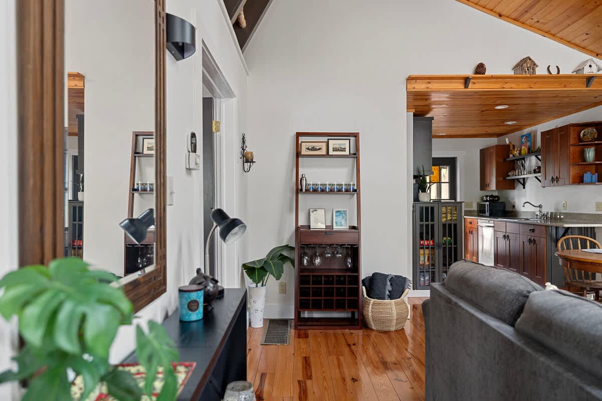A bright living room features a stone fireplace, a gray sectional sofa, and a round wooden dining table, all illuminated by a skylight and a modern pendant light under a vaulted wooden ceiling.