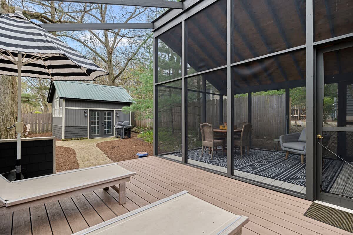 Expansive wooden deck with lounge chairs and a striped umbrella, adjacent to a screened porch featuring a dining table and chairs. The dark grey shed is visible in the background.