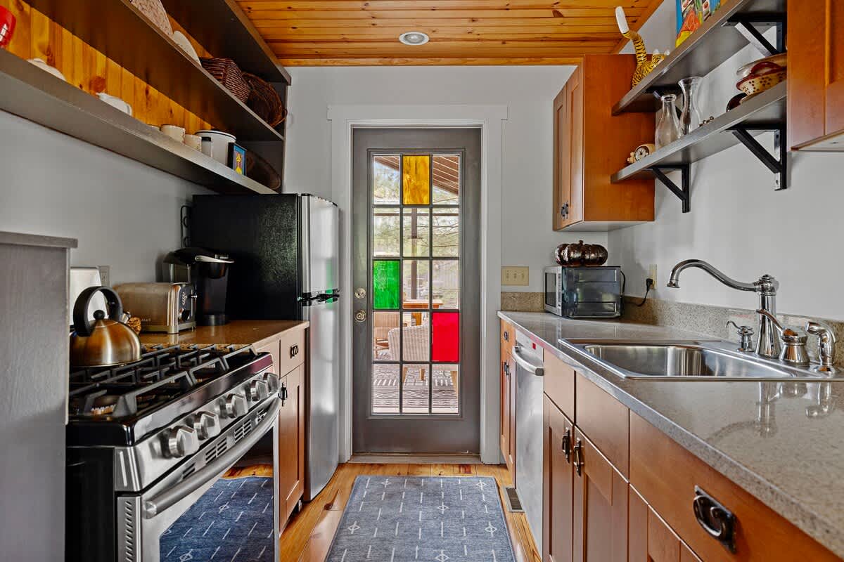 Narrow galley kitchen featuring a stainless steel gas range, refrigerator, microwave, and wooden cabinetry, with a colorful stained glass door at the far end leading to an outdoor area.