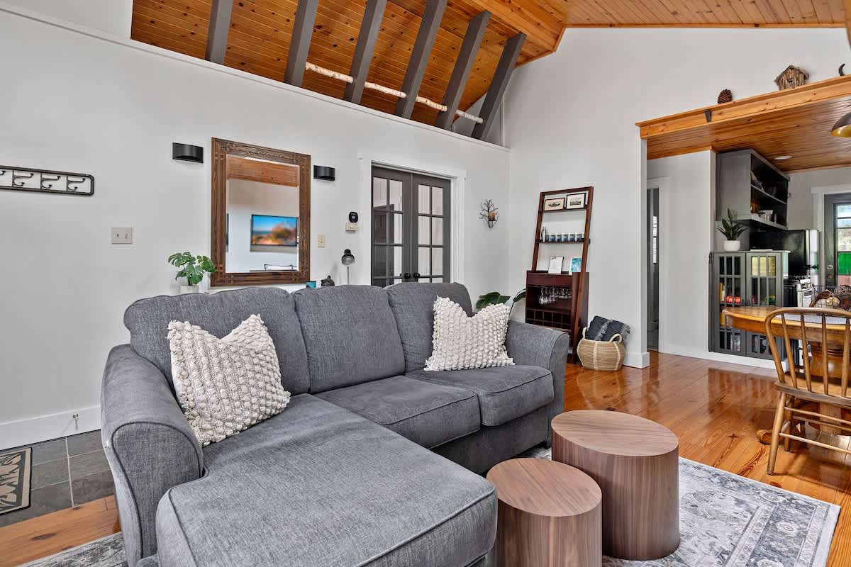 A wide view of the living room showcases a gray sectional sofa with wooden coffee tables, leading into the kitchen area, featuring a vaulted wooden ceiling and polished hardwood floors.