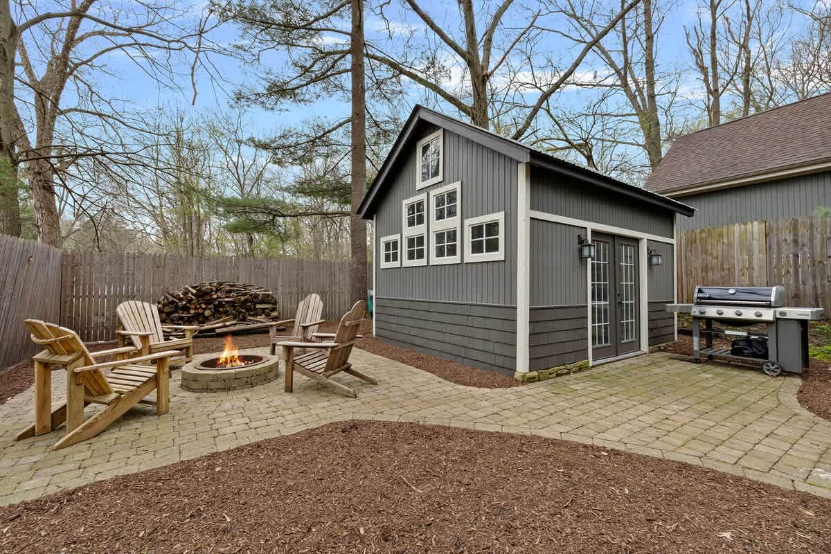 Outdoor fire pit area with four wooden Adirondack chairs surrounding a stone fire pit, set on a paver patio. A dark grey shed with white trim and a grill are visible in the background.