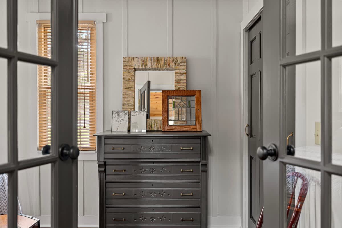 Dark wooden dresser adorned with decorative mirrors and picture frames, positioned between two dark-framed French doors, with light paneled walls and wooden blinds on a window.