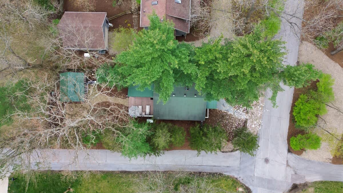 Aerial view of the property showcasing a dark gray house with a green roof, nestled among lush green trees. The surrounding neighborhood with other homes and roads is visible.