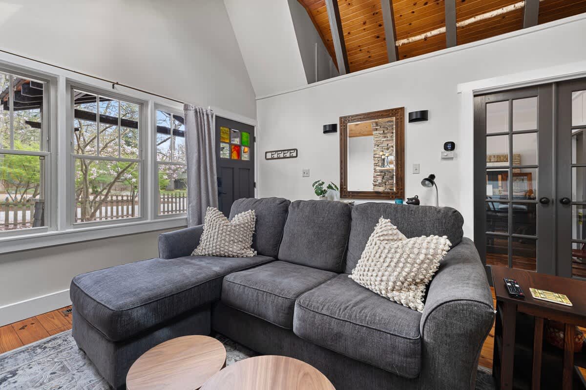 A spacious living room is furnished with a gray sectional sofa, a dark front door featuring colorful stained glass, and large windows, all beneath a vaulted wooden ceiling.
