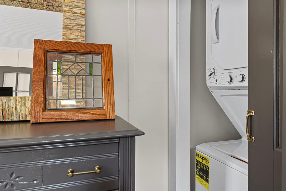 A stacked white washer and dryer unit is visible next to a dark dresser with a decorative stained glass piece. The area features light walls and a simple trim.