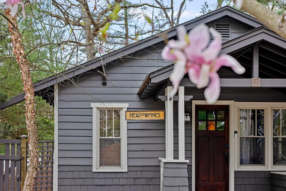 A slightly angled view of the dark gray cottage's front entrance, highlighting the covered porch with white pillars and a dark wood door, framed by a blurred pink magnolia blossom in the foreground. The house features horizontal siding on the upper half and shingle siding below, with a 'MEADOWPINES' sign.