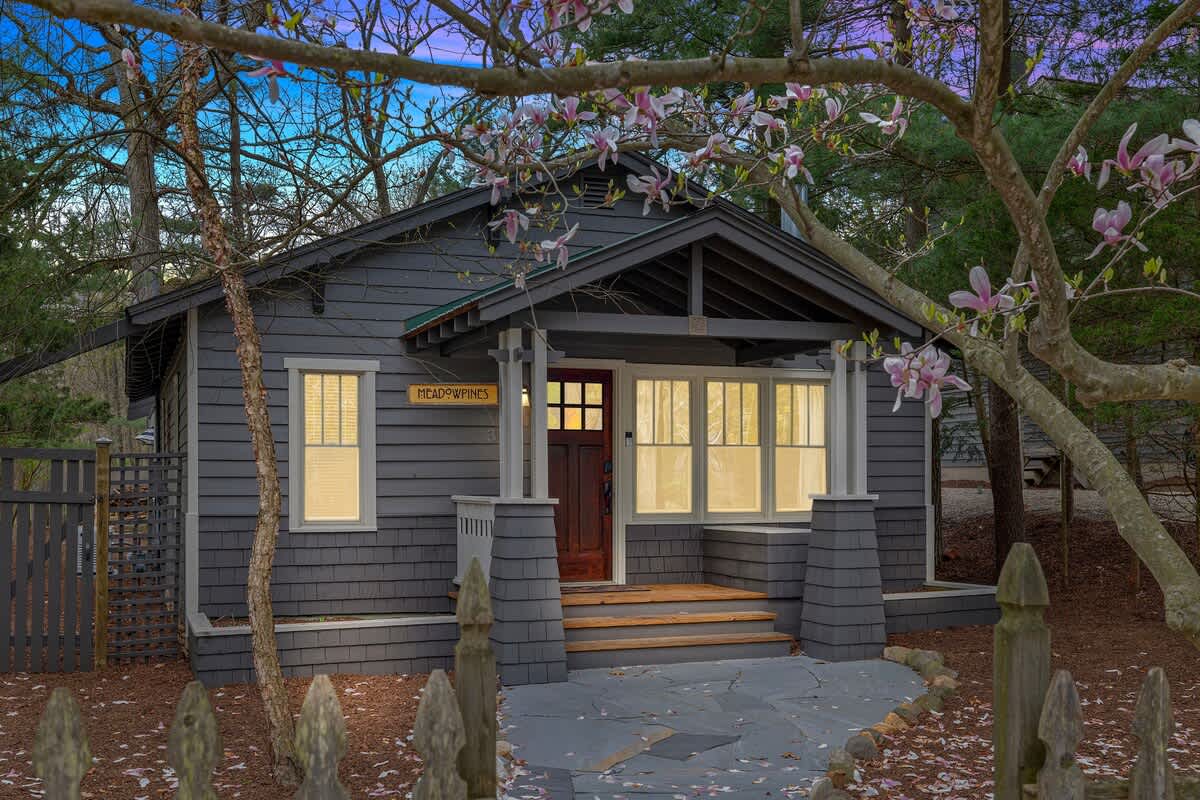 Charming grey bungalow-style home with a welcoming front porch, dark wood door, and white trim windows. Blooming magnolia trees frame the entrance, and a small picket fence lines the path.