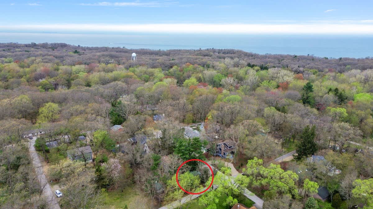 Wide aerial view showing the property, circled in red, nestled within a vast forest landscape. A large body of water, likely Lake Michigan, is visible in the distant background under a blue sky.