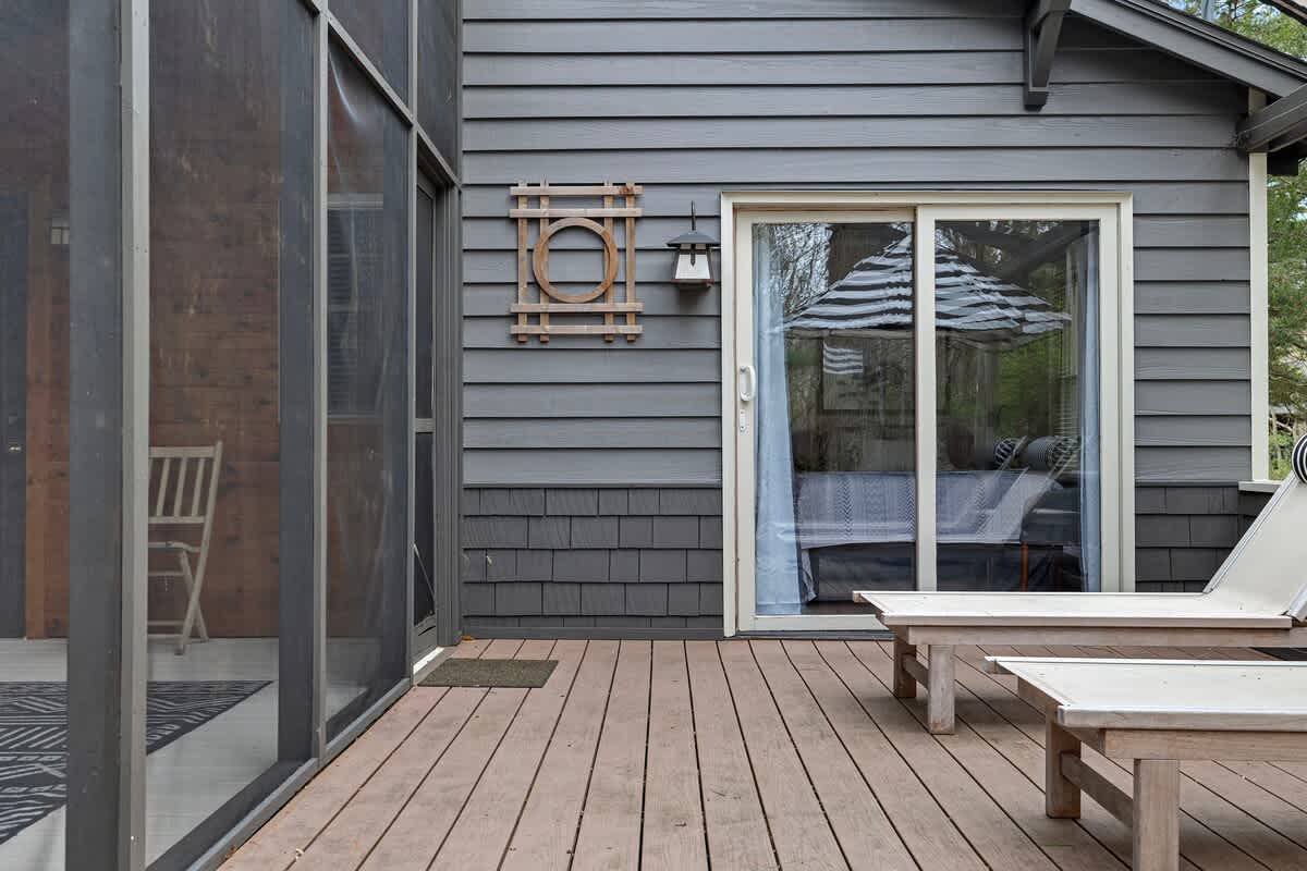Exterior view of the house's back, featuring a large sliding glass door leading to a wooden deck with lounge chairs. A screened porch is visible to the left, and the house has dark grey siding.