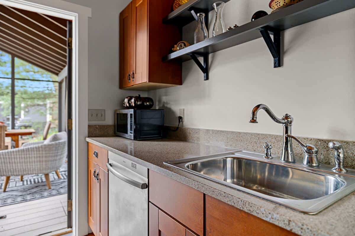 Detailed view of the kitchen sink area with a stainless steel sink, modern faucet, dishwasher, and a microwave on the counter, with a screened porch visible through the open doorway.