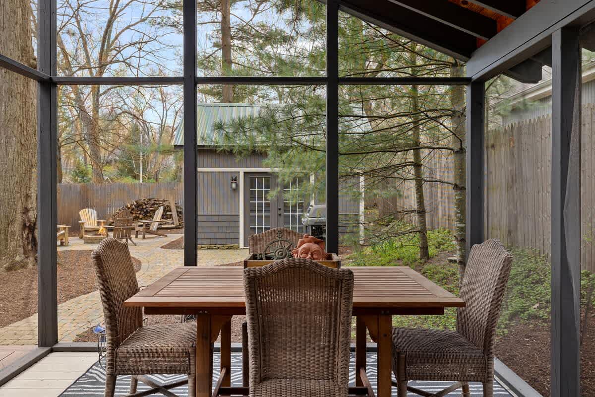 View from a screened porch showing a wooden dining table with four wicker chairs. Beyond the screen, a paved patio with Adirondack chairs and a fire pit is visible, along with a gray shed.