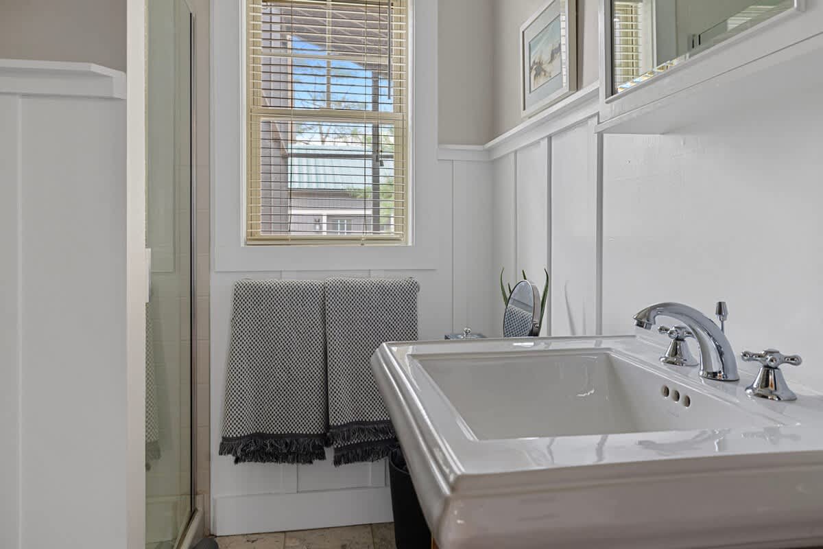 Close-up of the bright bathroom's white pedestal sink with chrome fixtures, next to a glass-enclosed shower. A window with blinds and wainscoting are visible.