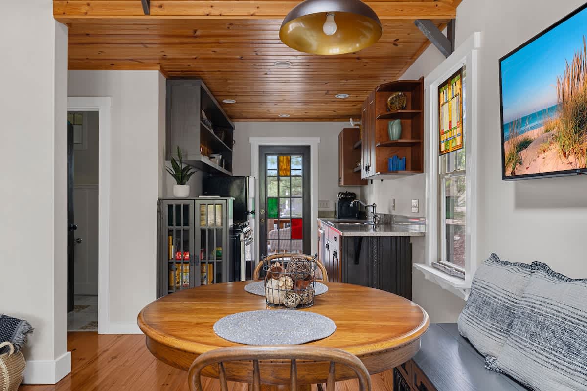 View of the dining area with a round wooden table and bench seating, extending into a compact kitchen with dark cabinetry and a stained glass door, all beneath a warm wooden ceiling.