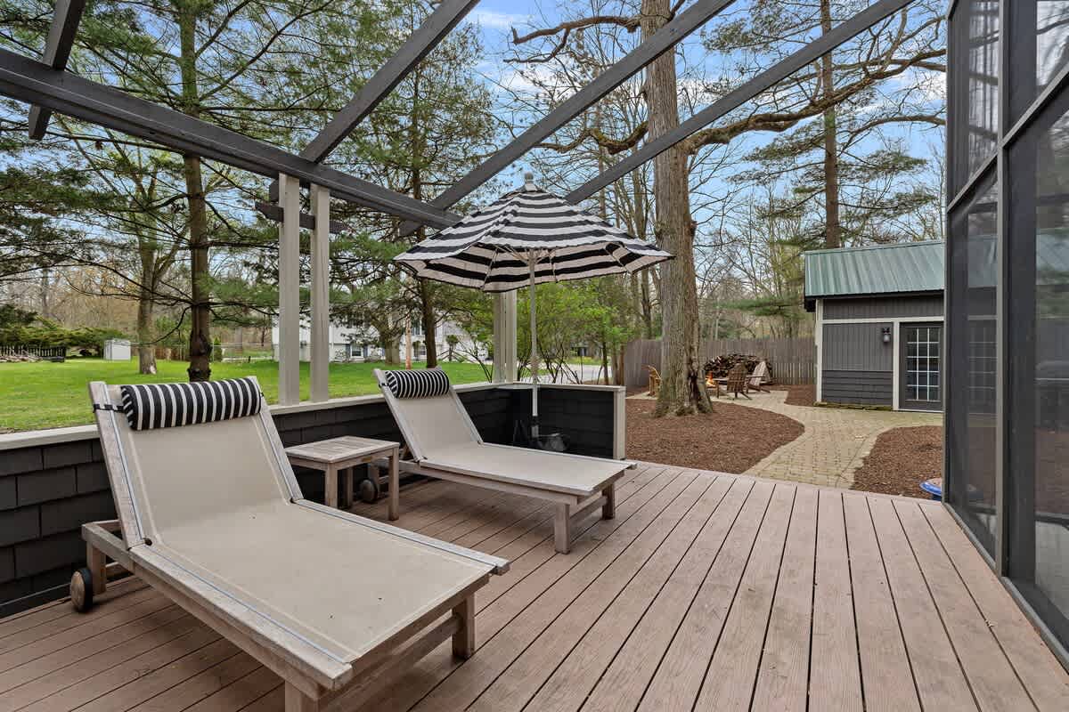 Spacious outdoor deck with two light-colored lounge chairs and a striped umbrella, overlooking a grassy yard. The dark grey shed and fire pit area are visible in the distance.