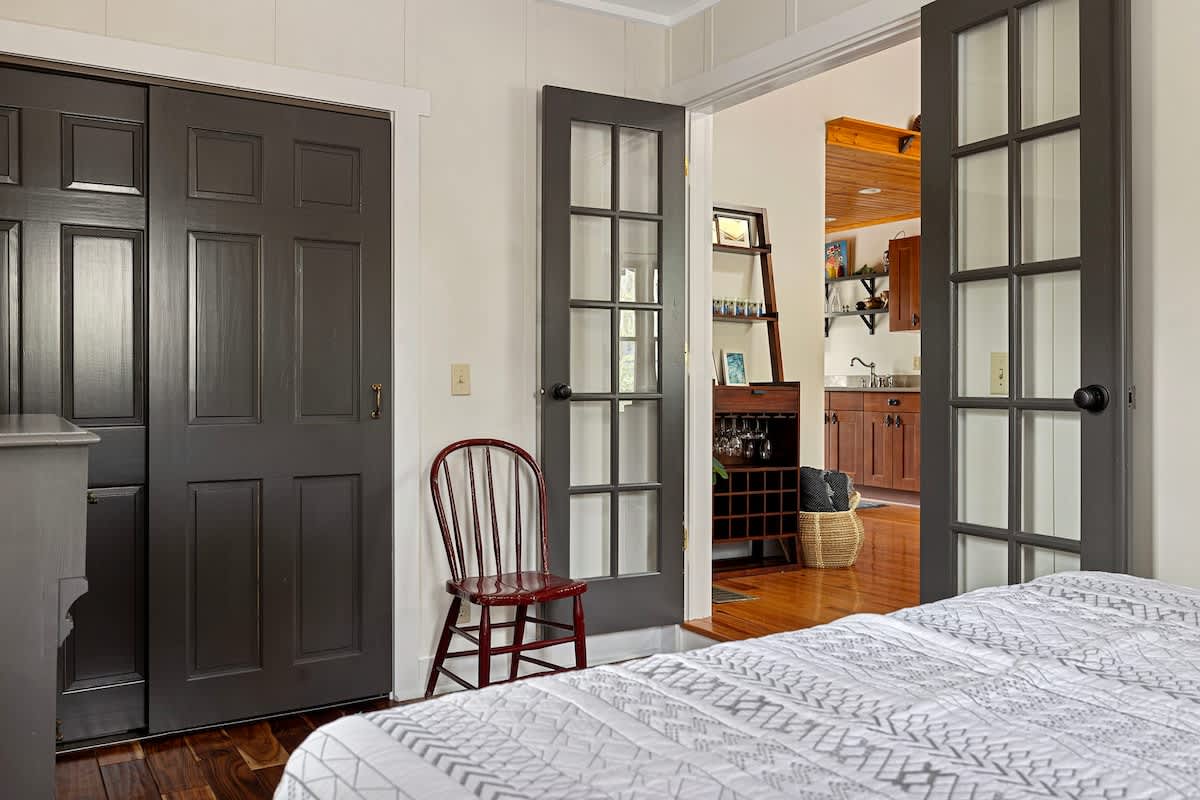 View from a bedroom looking out through dark French doors into the main living area, featuring a dark wooden closet, a red accent chair, and hardwood floors.