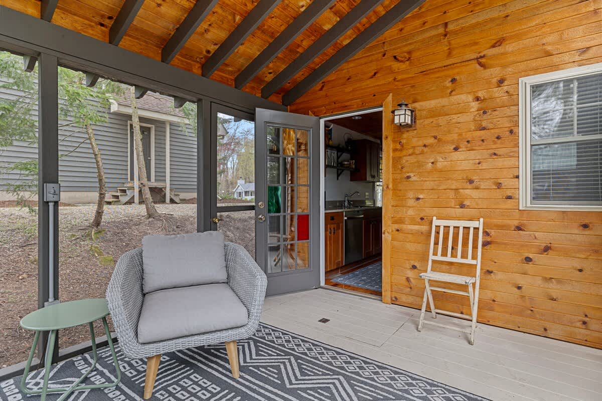 Cozy screened porch with a grey wicker armchair, a small green side table, and a patterned rug. A glass-paned door opens into an interior space with wood paneling and a small kitchen area.