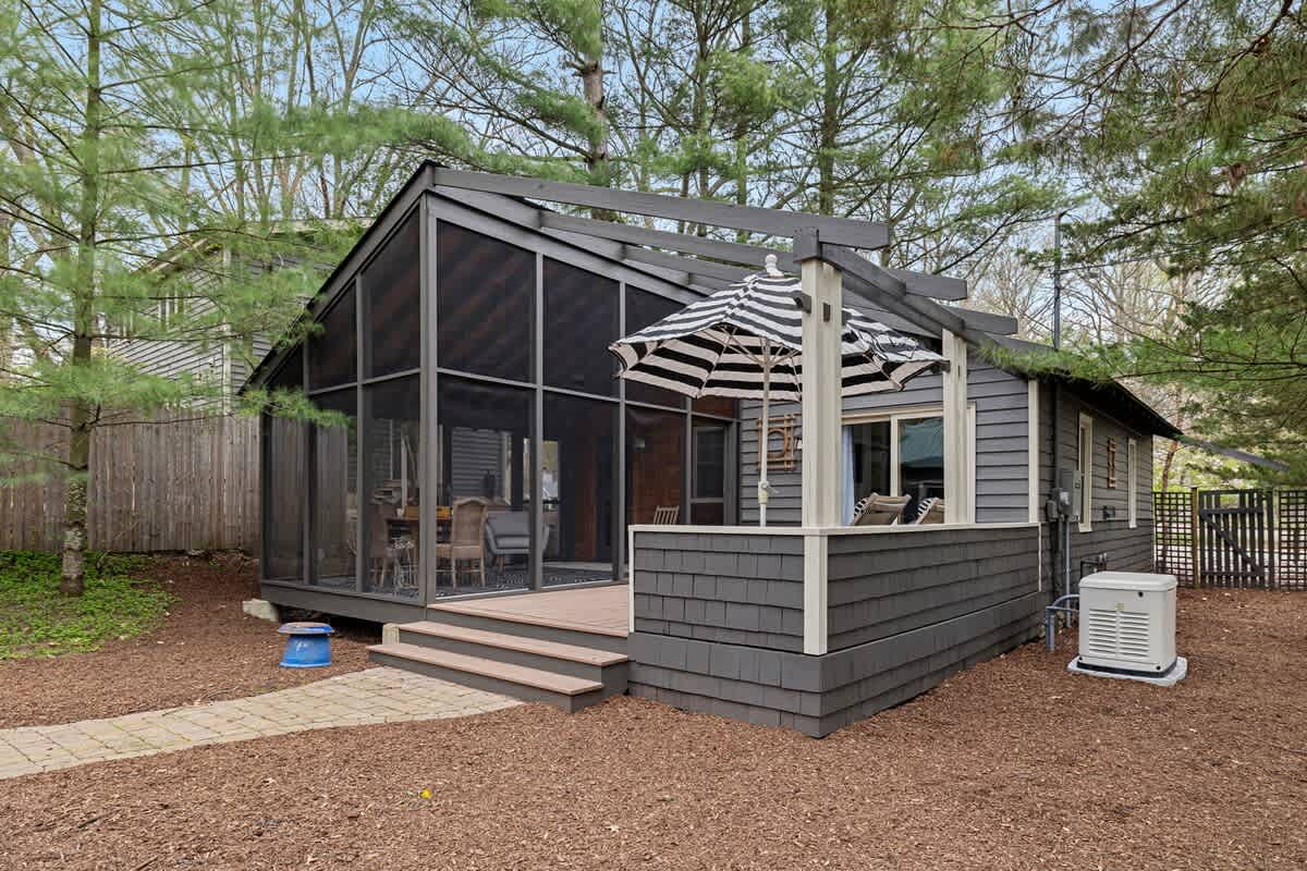 Dark gray house with shingle siding features a screened porch and an open deck with a black and white striped umbrella. The surrounding ground is covered in brown mulch with a stone path.