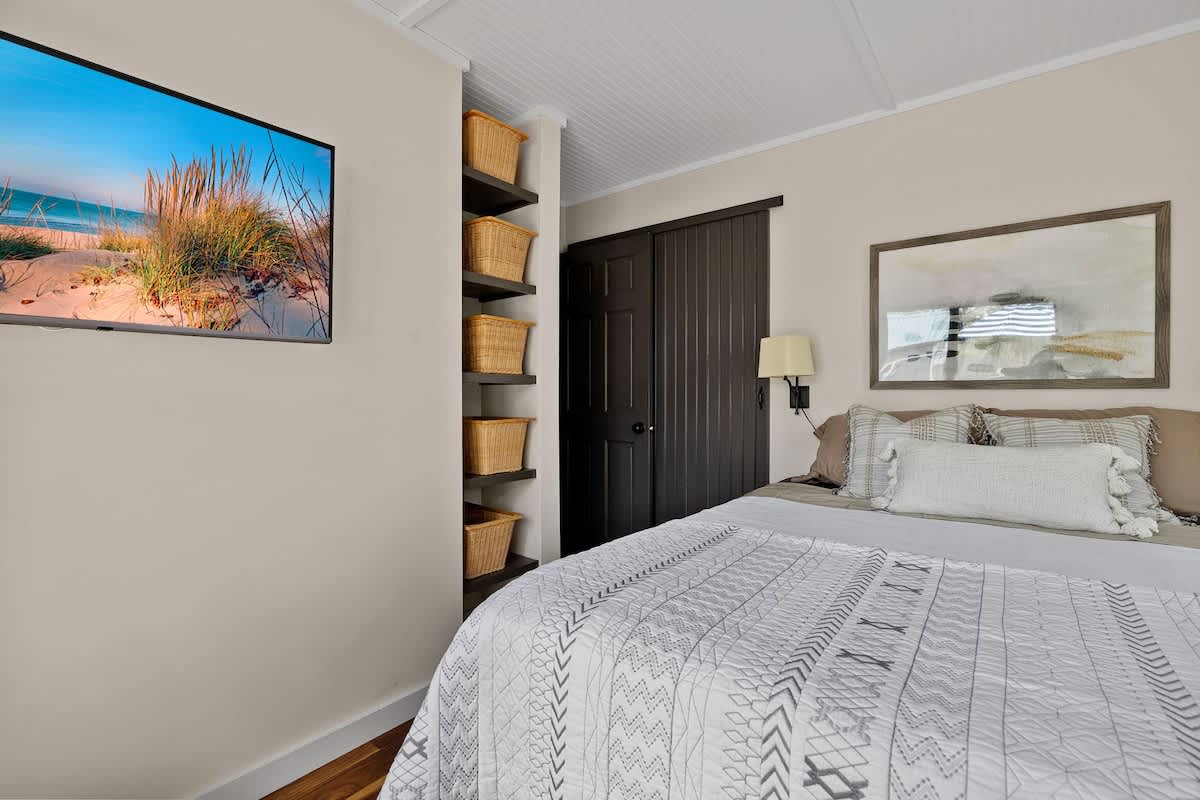 Bright bedroom featuring a queen-sized bed with a white and gray patterned quilt, a wall-mounted TV, and built-in shelving with woven baskets, next to a dark wooden closet.